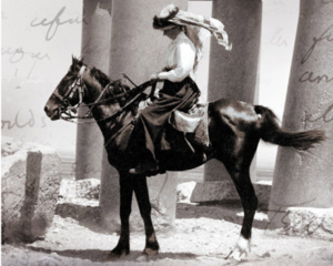 Black-and-white archival photograph of a woman in a wide-brimmed hat and long skirt riding a dark horse among tall stone columns, with handwritten script layered in the background.