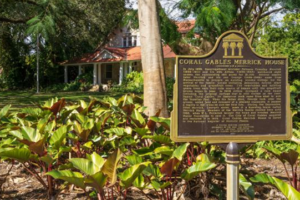 Historic marker for the Coral Gables Merrick House stands among tropical plants, with the red-roofed house visible through trees in the background.