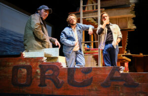 Three actors stand on a stage set designed as the damaged boat Orca in The Shark Is Broken.