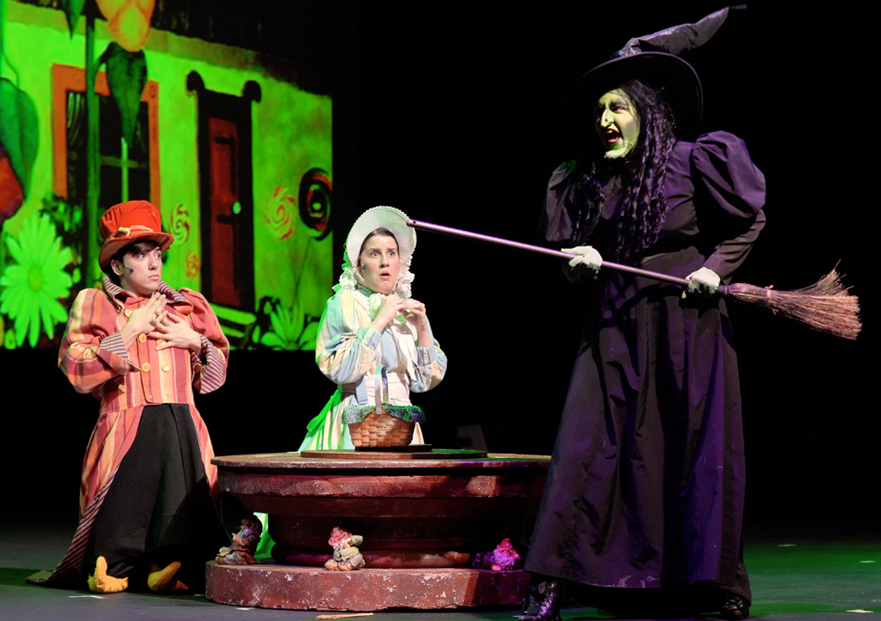 Stage scene from The Wizard of Oz showing the Wicked Witch holding a broom beside two costumed characters near a well.