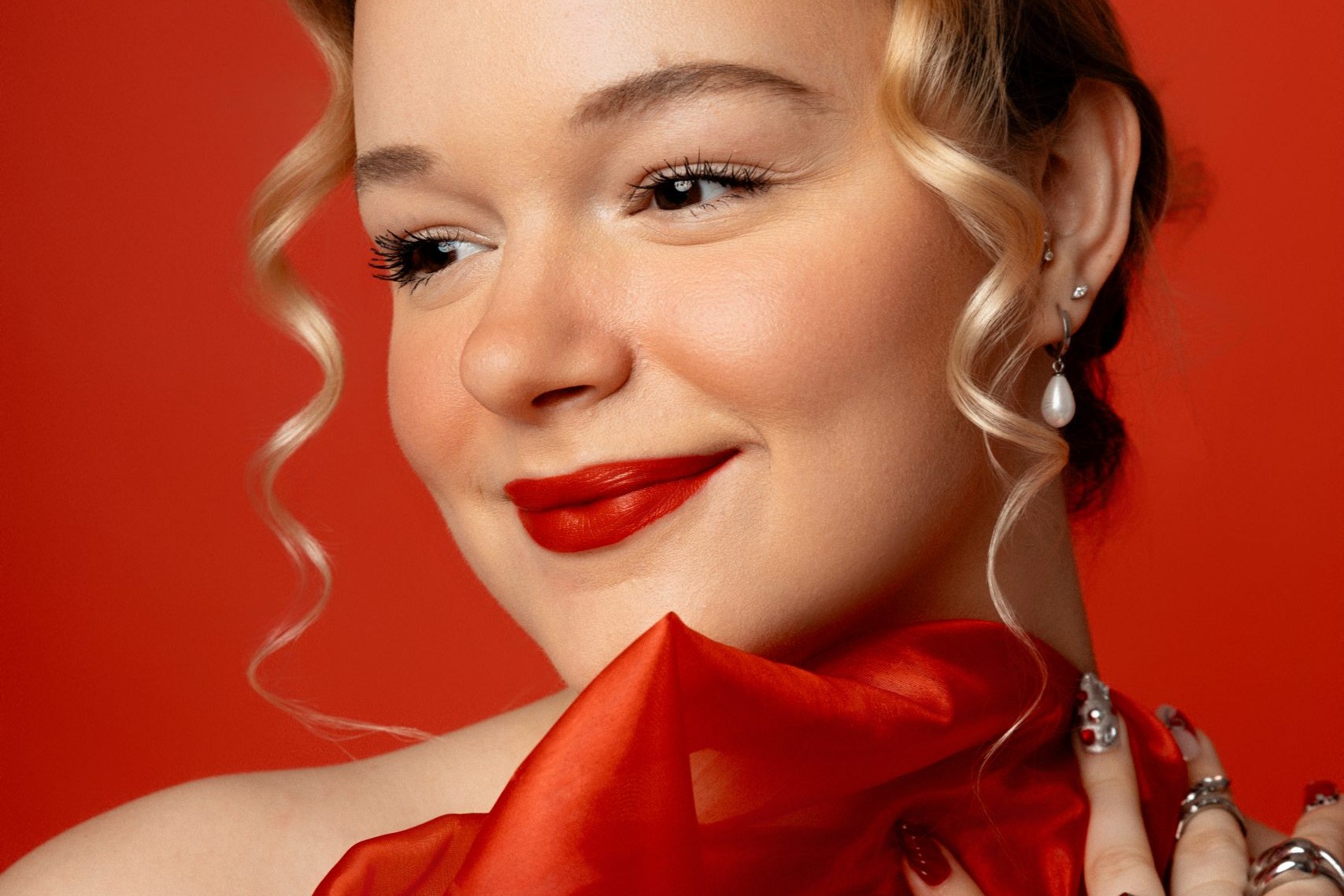 Close-up portrait of a blonde woman wearing red lipstick and holding red fabric against a vivid red background.