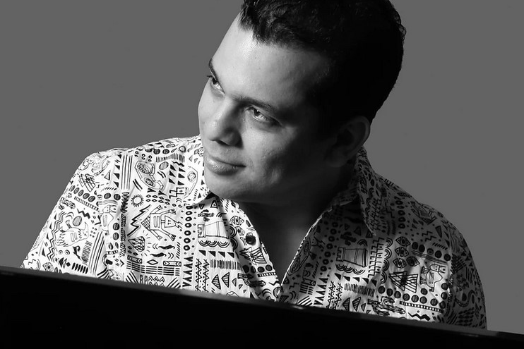 Black-and-white portrait of César Orozco seated at a piano, wearing a patterned shirt and looking off to the side.