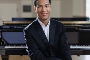 Smiling pianist in a dark suit and white shirt seated in front of a grand piano.