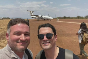 Henry Parra, left, and Adam Hlavaty take a selfie on an airfield in Kenya, with a small propeller aircraft on the runway behind them.