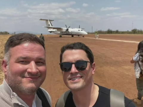 Henry Parra, left, and Adam Hlavaty take a selfie on an airfield in Kenya, with a small propeller aircraft on the runway behind them.