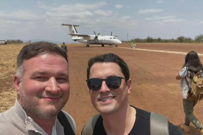 Henry Parra, left, and Adam Hlavaty take a selfie on an airfield in Kenya, with a small propeller aircraft on the runway behind them.