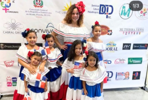 An adult and six children pose in white, red and blue Dominican-style outfits, some holding or wearing Dominican Republic flag colors, in front of a sponsor backdrop.