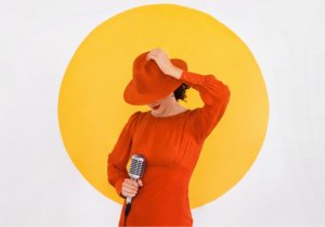 Jazz vocalist Laila Biali in a red dress and wide-brimmed red hat holding a vintage-style microphone in front of a large yellow circle.