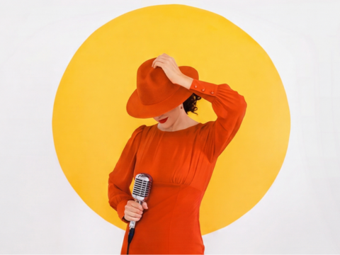 Jazz vocalist Laila Biali in a red dress and wide-brimmed red hat holding a vintage-style microphone in front of a large yellow circle.
