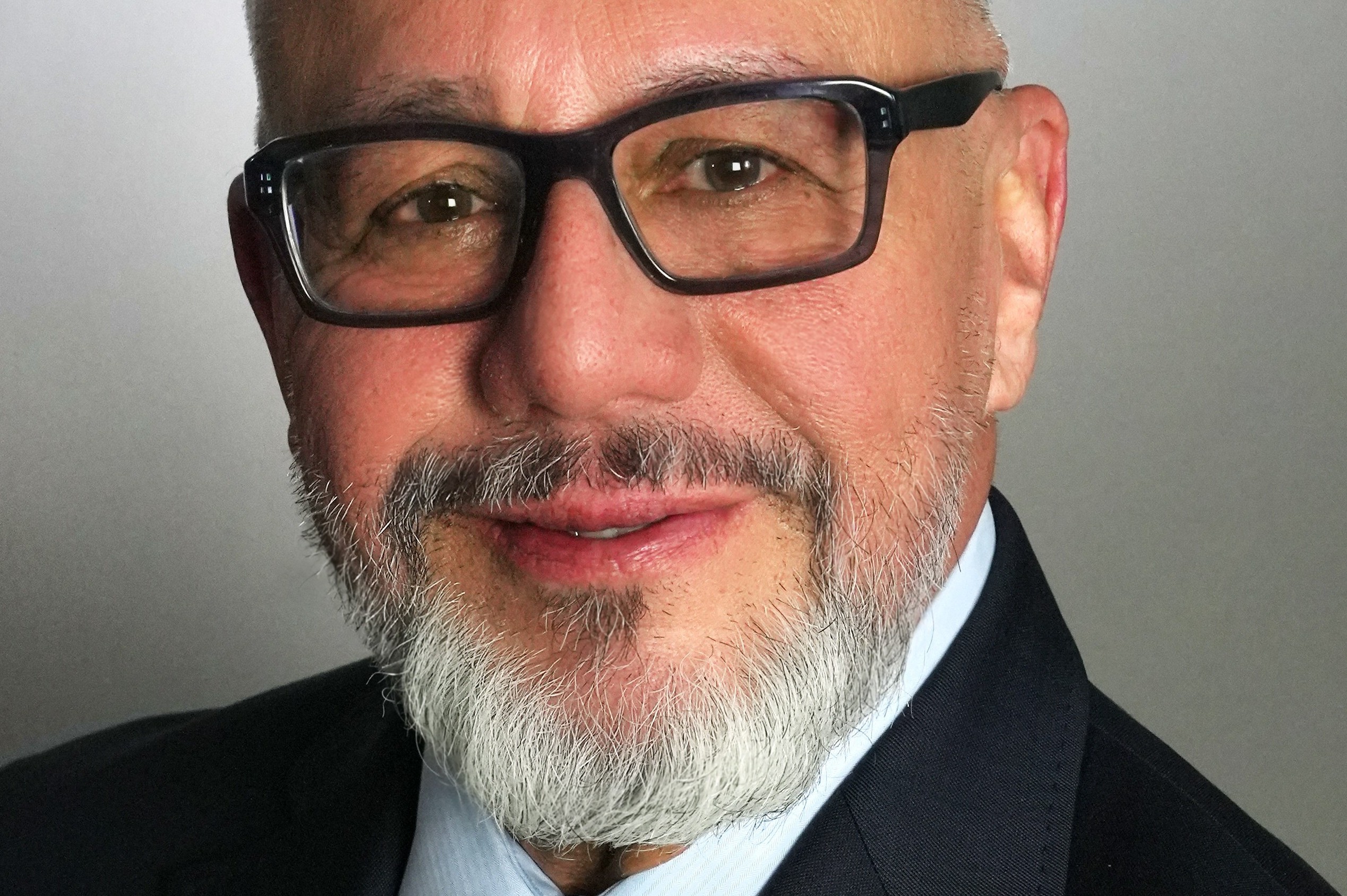 Close-up portrait of Joe L. Gómez wearing dark rectangular glasses and a suit, with a gray beard against a plain gray background.