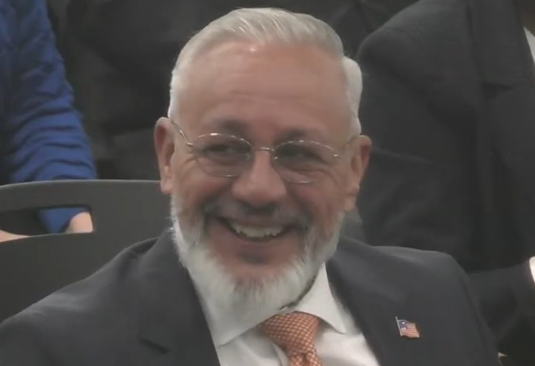 Joe L. Gómez, wearing a dark suit and orange tie, smiles while seated during a public meeting.