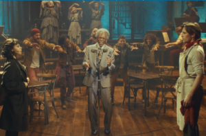 Performers on the Hadestown stage surround a gray-haired man singing at a vintage microphone, with wooden tables and chairs and an industrial-style set behind them.