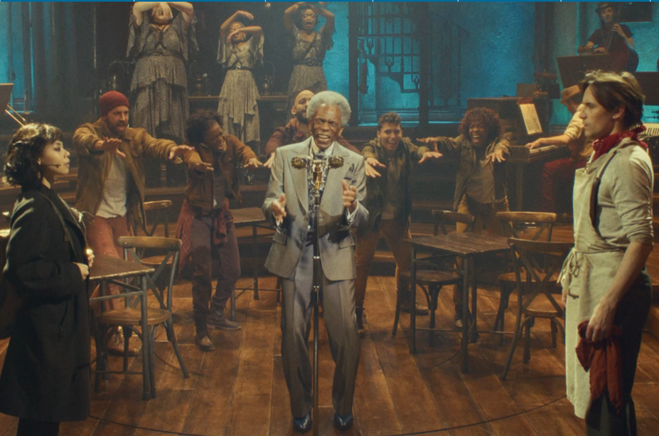 Performers on the Hadestown stage surround a gray-haired man singing at a vintage microphone, with wooden tables and chairs and an industrial-style set behind them.