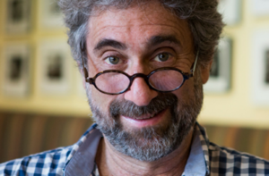 Close-up portrait of a smiling man with curly gray hair, a gray beard and round glasses, wearing a blue-and-white checkered shirt.