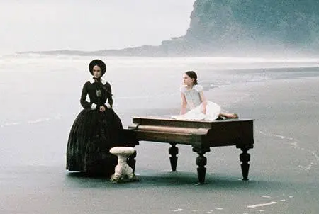 A woman in a long black dress and a young girl in white stand beside a grand piano on a misty beach.