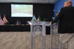 A man speaks at a lectern before the Coral Gables City Commission as a presentation reading “Stop The Lies, Save The Garden” appears on a screen behind the dais.
