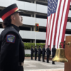 Coral Gables firefighter standing beside a brass ceremonial bell, with an honor guard and a large American flag in the background during a 9/11 remembrance ceremony.