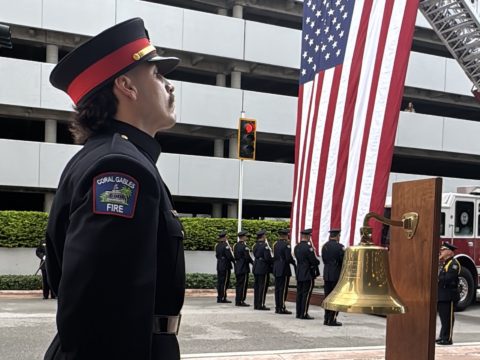 Coral Gables firefighter standing beside a brass ceremonial bell, with an honor guard and a large American flag in the background during a 9/11 remembrance ceremony.