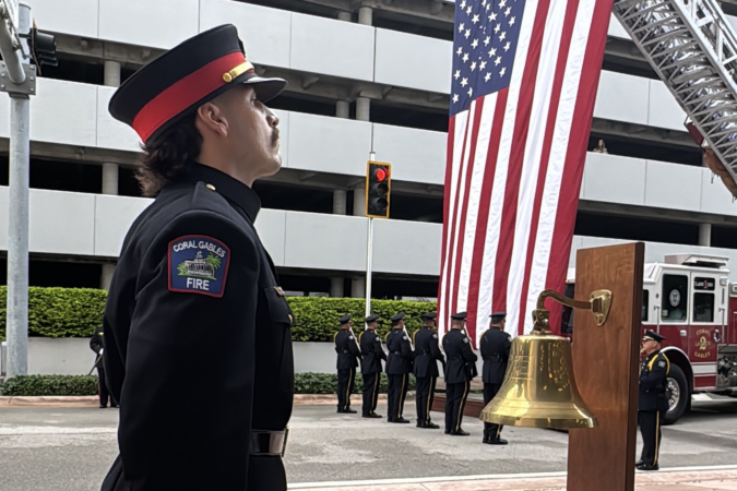 Coral Gables firefighter standing beside a brass ceremonial bell, with an honor guard and a large American flag in the background during a 9/11 remembrance ceremony.