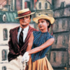 A man and woman in period clothing and straw hats pose together against a painted Paris street backdrop in a scene from An American in Paris.