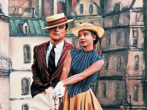 A man and woman in period clothing and straw hats pose together against a painted Paris street backdrop in a scene from An American in Paris.