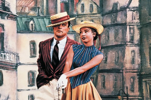 A man and woman in period clothing and straw hats pose together against a painted Paris street backdrop in a scene from An American in Paris.