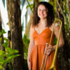 Allie Biancoviso in an orange dress stands among tropical plants holding a brass trombone.
