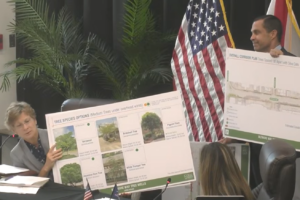 Vice Mayor Rhonda Anderson and Mayor Vince Lago hold large presentation boards showing tree species and corridor plans during a Coral Gables City Commission meeting.