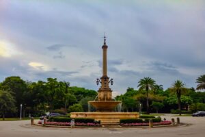 DeSoto Fountain stands at the center of a landscaped traffic circle in Coral Gables, surrounded by palm trees, roadway and passing cars.