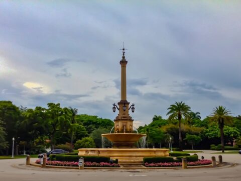 DeSoto Fountain stands at the center of a landscaped traffic circle in Coral Gables, surrounded by palm trees, roadway and passing cars.