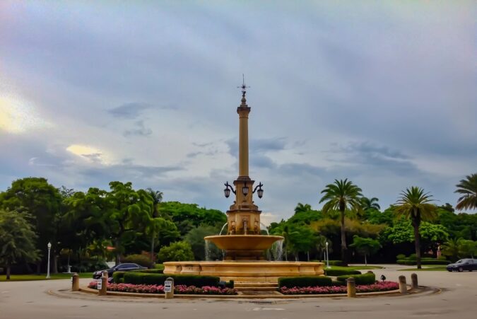 DeSoto Fountain stands at the center of a landscaped traffic circle in Coral Gables, surrounded by palm trees, roadway and passing cars.