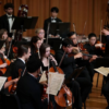 Conductor Gerard Schwarz leads the Frost Symphony Orchestra in performance, with string and woodwind players seated onstage.
