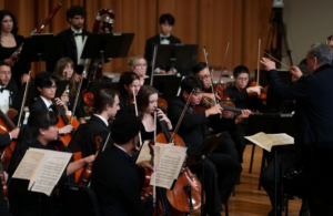 Conductor Gerard Schwarz leads the Frost Symphony Orchestra in performance, with string and woodwind players seated onstage.