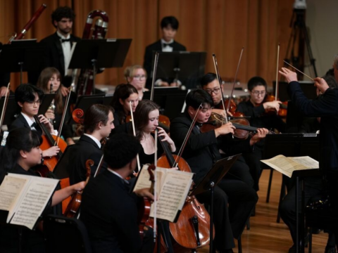 Conductor Gerard Schwarz leads the Frost Symphony Orchestra in performance, with string and woodwind players seated onstage.