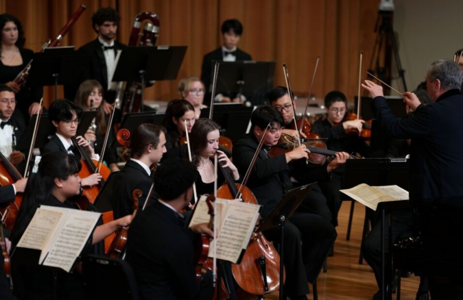 Conductor Gerard Schwarz leads the Frost Symphony Orchestra in performance, with string and woodwind players seated onstage.