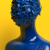 Blue sculptural human figure with floral and botanical forms covering the head, set against a bright yellow background.