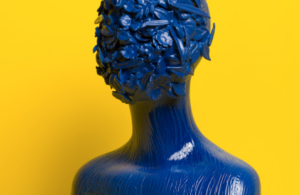 Blue sculptural human figure with floral and botanical forms covering the head, set against a bright yellow background.