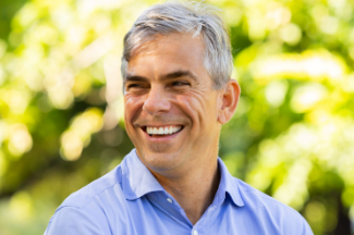 Portrait of Nestor Menendez smiling outdoors in a light blue collared shirt with trees blurred in the background.