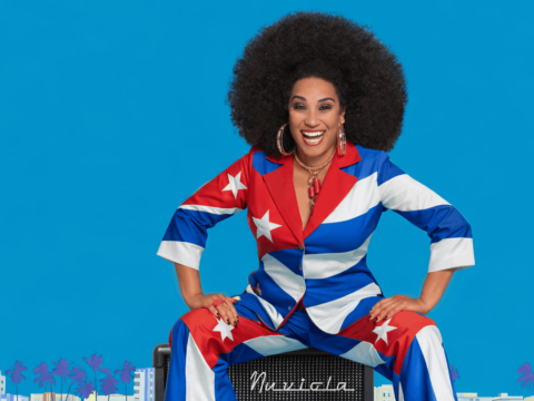 Smiling Cuban vocalist in a red, white and blue suit inspired by the Cuban flag poses against a bright blue background.