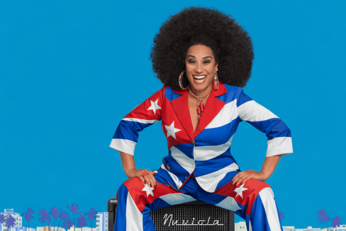 Smiling Cuban vocalist in a red, white and blue suit inspired by the Cuban flag poses against a bright blue background.