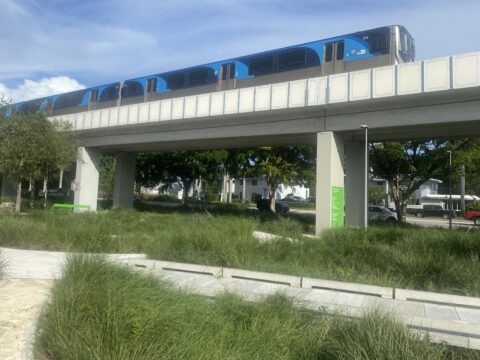 Blue Metrorail train travels on an elevated guideway above landscaped paths and greenery along The Underline in Coral Gables.