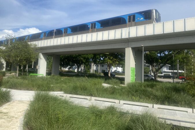 Blue Metrorail train travels on an elevated guideway above landscaped paths and greenery along The Underline in Coral Gables.