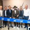 Eight executives and civic leaders stand behind a blue BMI ribbon at the opening of the music licensing company's Coral Gables office. A man in a black hat at the center holds ceremonial scissors.
