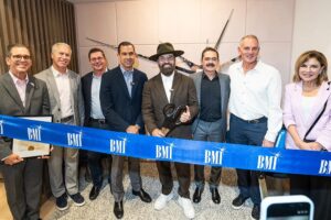 Eight executives and civic leaders stand behind a blue BMI ribbon at the opening of the music licensing company's Coral Gables office. A man in a black hat at the center holds ceremonial scissors.
