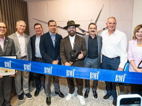 Eight executives and civic leaders stand behind a blue BMI ribbon at the opening of the music licensing company's Coral Gables office. A man in a black hat at the center holds ceremonial scissors.