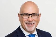 Headshot of Genaro “Chip” Iglesias wearing black-framed glasses, a dark suit, white shirt and blue tie, smiling against a light background.