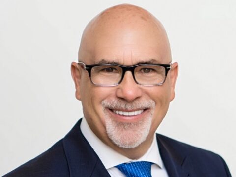 Headshot of Genaro “Chip” Iglesias wearing black-framed glasses, a dark suit, white shirt and blue tie, smiling against a light background.