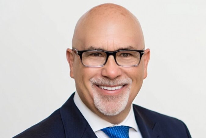 Headshot of Genaro “Chip” Iglesias wearing black-framed glasses, a dark suit, white shirt and blue tie, smiling against a light background.