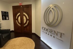 Interior of Cogent Bank’s Coral Gables office, with the bank’s logo mounted on a wall beside wooden double doors and a seating area.