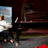 Two women in white blouses perform together at a black Steinway grand piano on a dimly lit stage, with an image of the Roman Colosseum behind them.